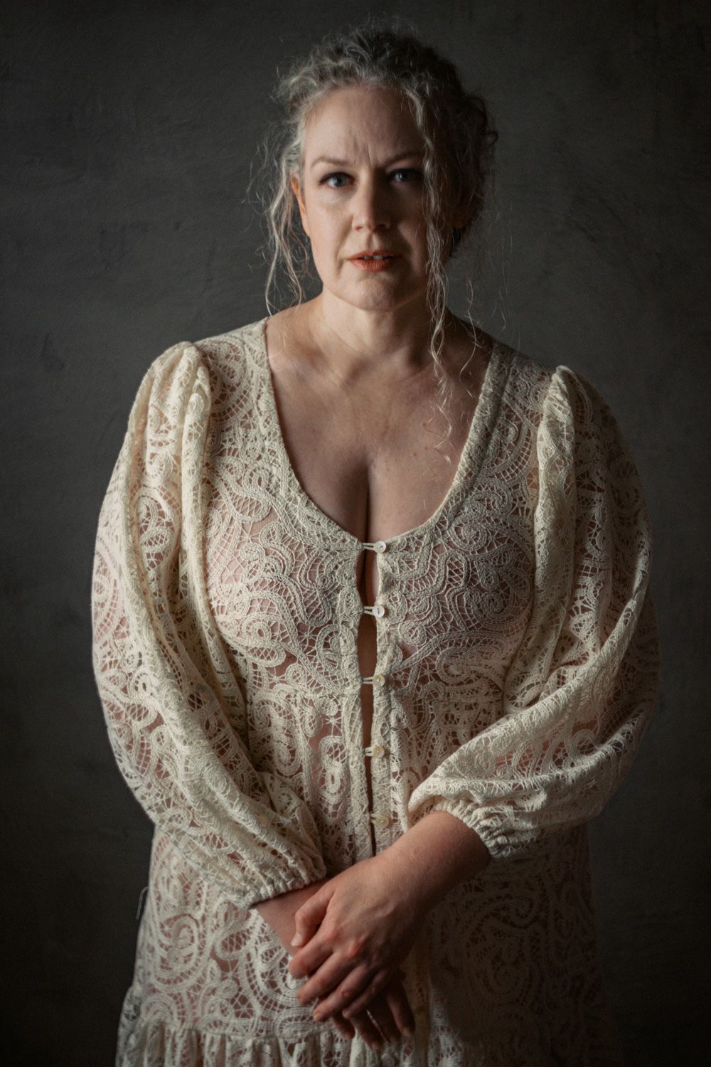A middle-aged woman with gray hair wearing an elegant cream lace dress poses against a dark gray studio background.