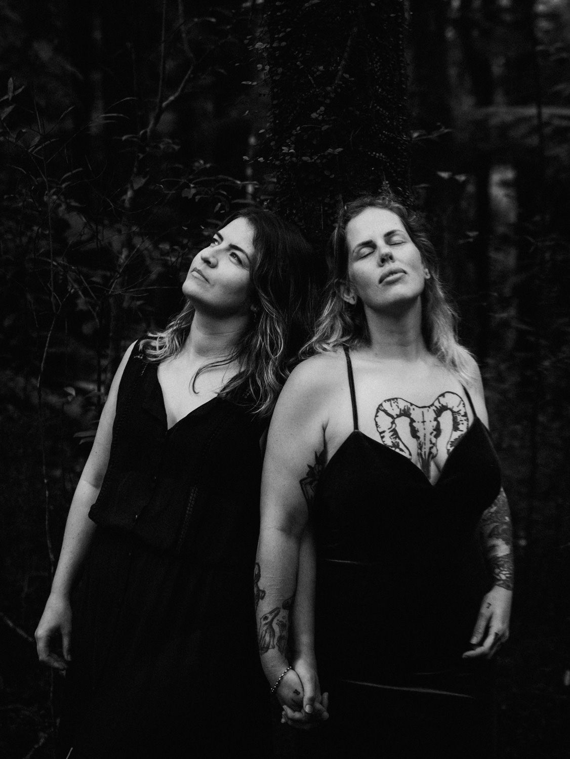 Two women in black dresses hold hands in a dark forest, one with tattoos, in a moody black and white portrait.