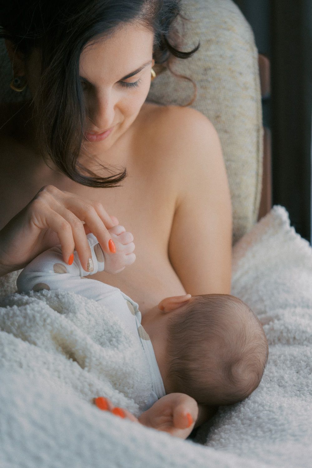 Mother breastfeeding newborn baby while sitting in a cozy chair covered with a soft white blanket.