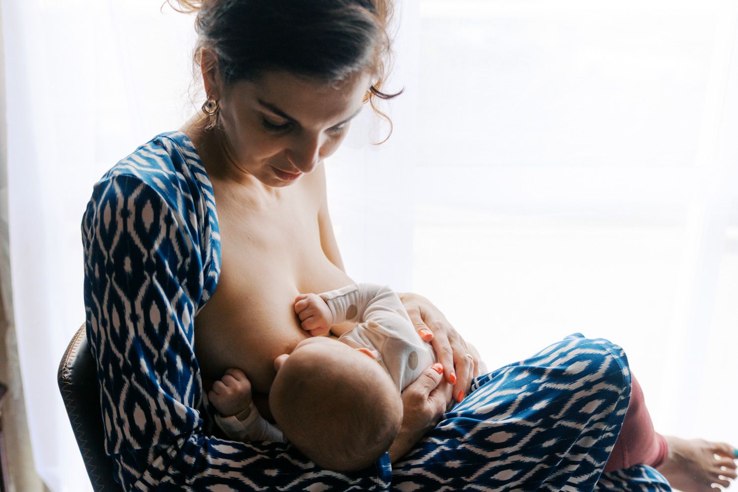 Mother breastfeeding her newborn baby while sitting in a chair near a bright window in a blue patterned wrap.