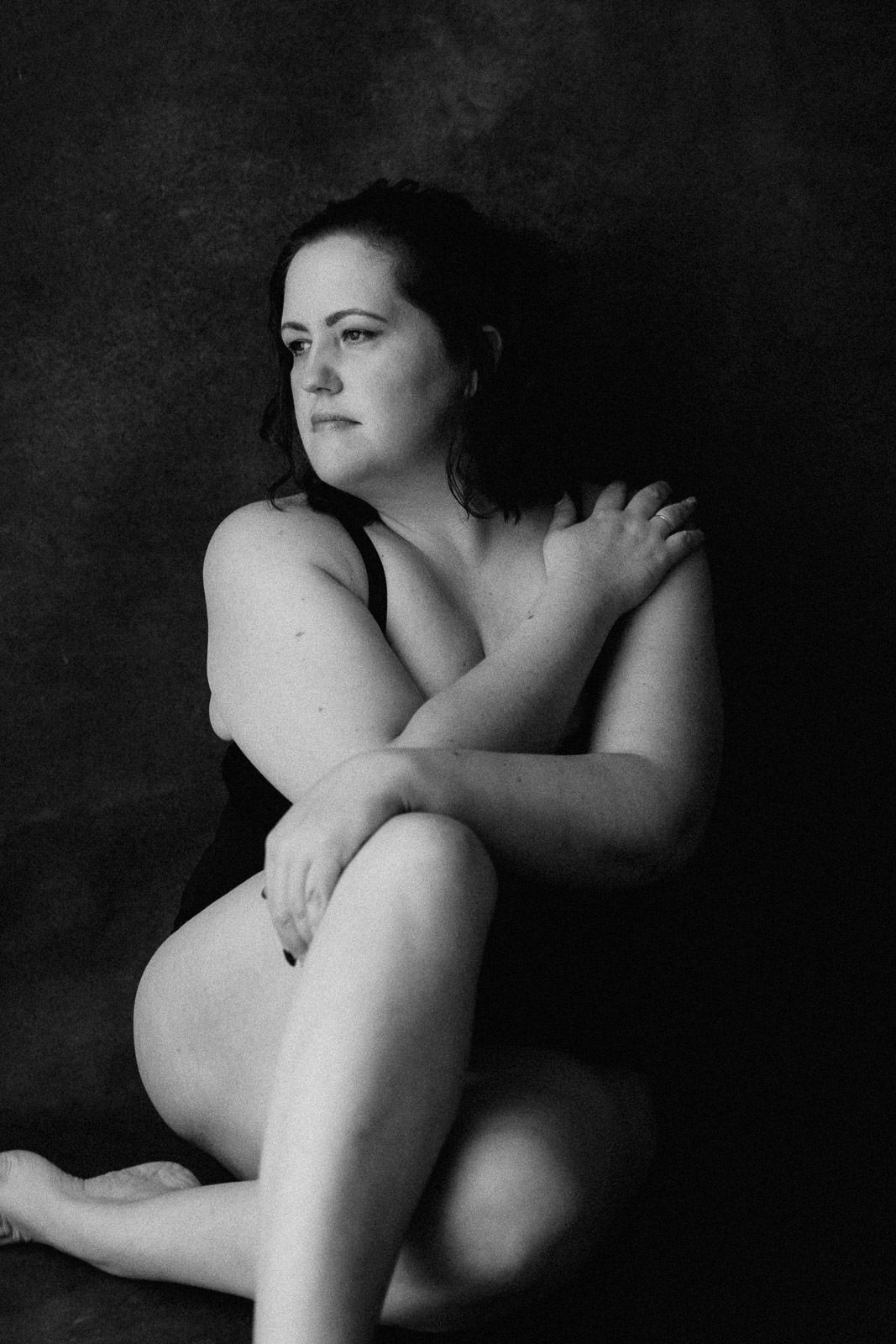 Black and white portrait of a woman sitting cross-legged, arms crossed over chest, gazing sideways against dark background.