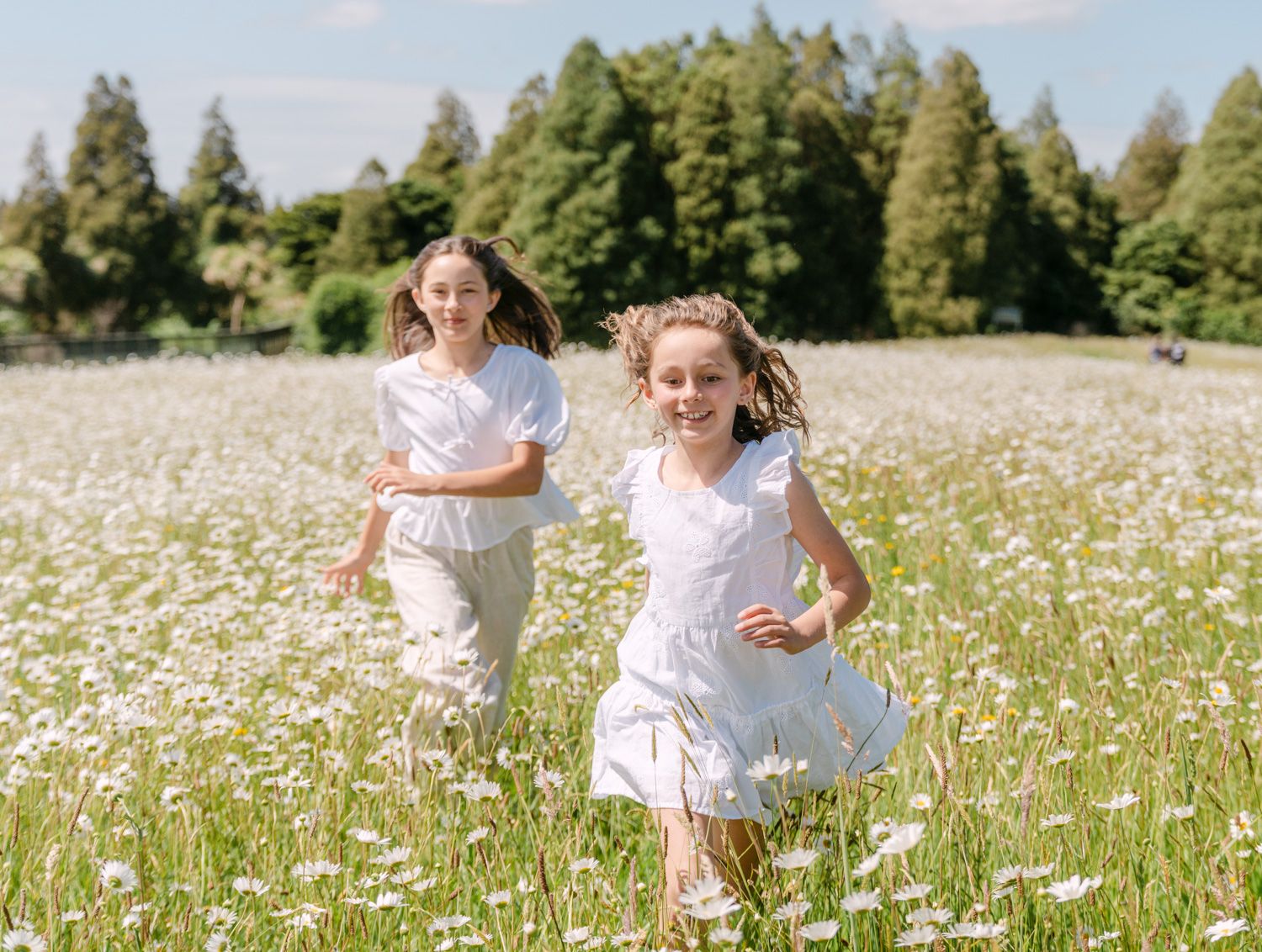 Two girls in white dresses running through a blooming daisy field with green trees in the background.