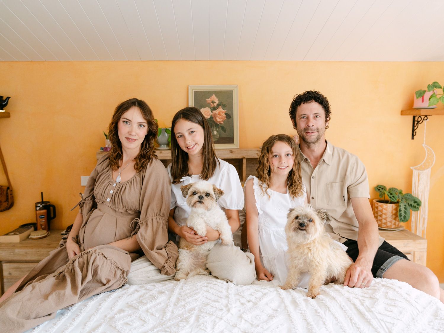 Family of four with two small dogs sitting together on a bed in a warmly decorated room.