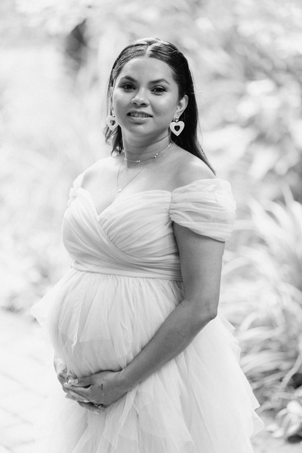 Black and white maternity photo of a smiling pregnant woman in a white off-shoulder tulle gown holding her baby bump.