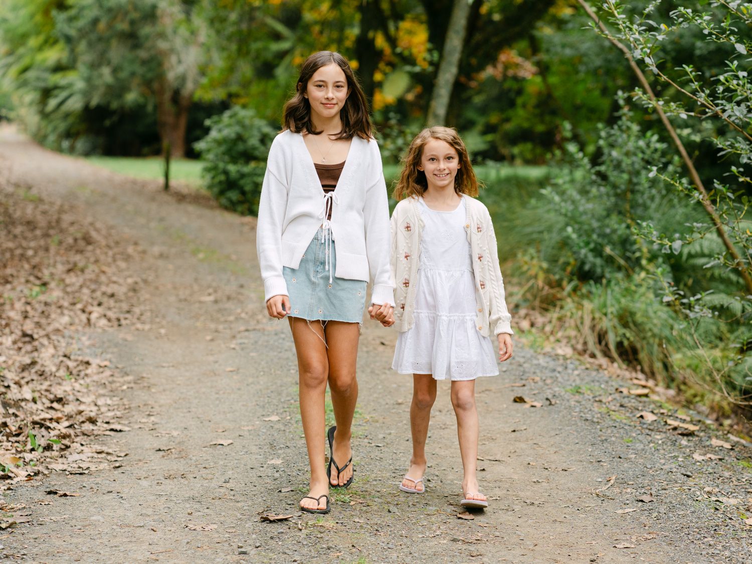 Two girls walking hand in hand on a garden path, wearing white outfits surrounded by lush green trees.