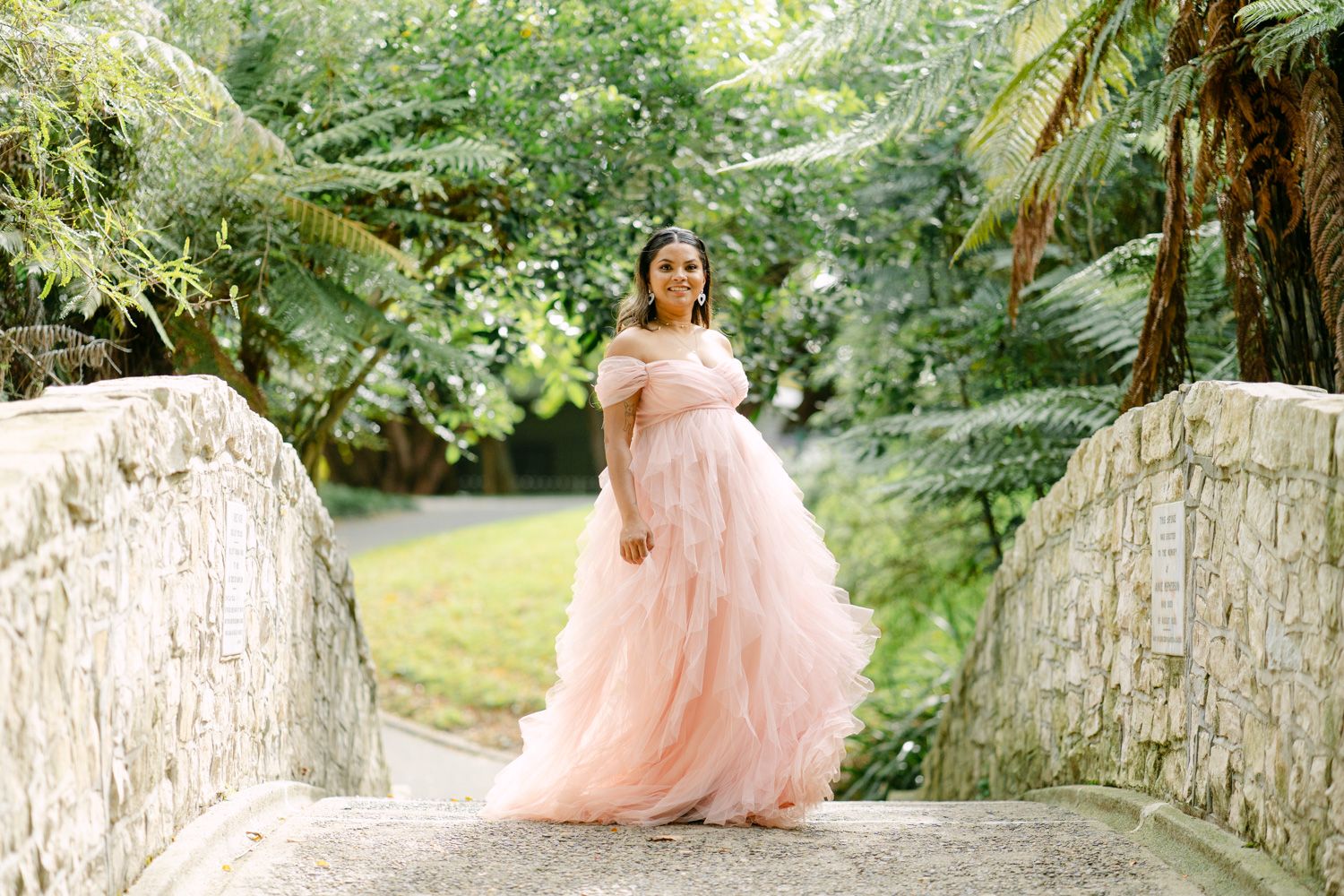 Woman in pink off-shoulder tulle gown poses on stone bridge surrounded by lush green tropical foliage.