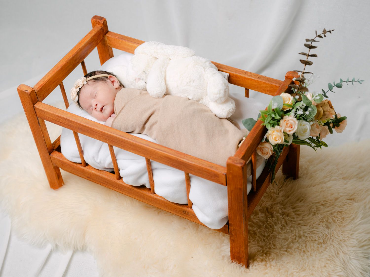 Newborn baby sleeping in a wooden crib with a white bunny toy and floral arrangement on a fluffy rug.