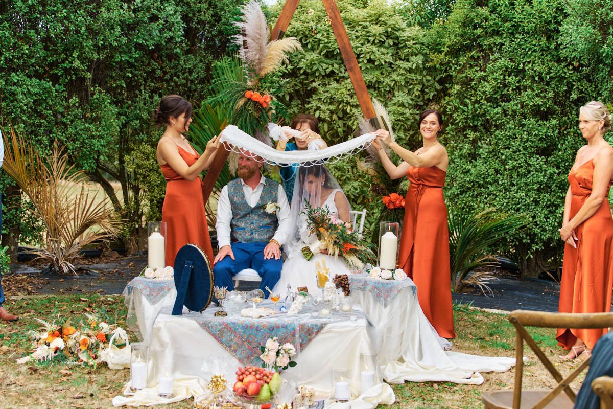 Outdoor wedding ceremony with couple under floral arch, bridesmaids in orange dresses, decorated table with candles and flowers.