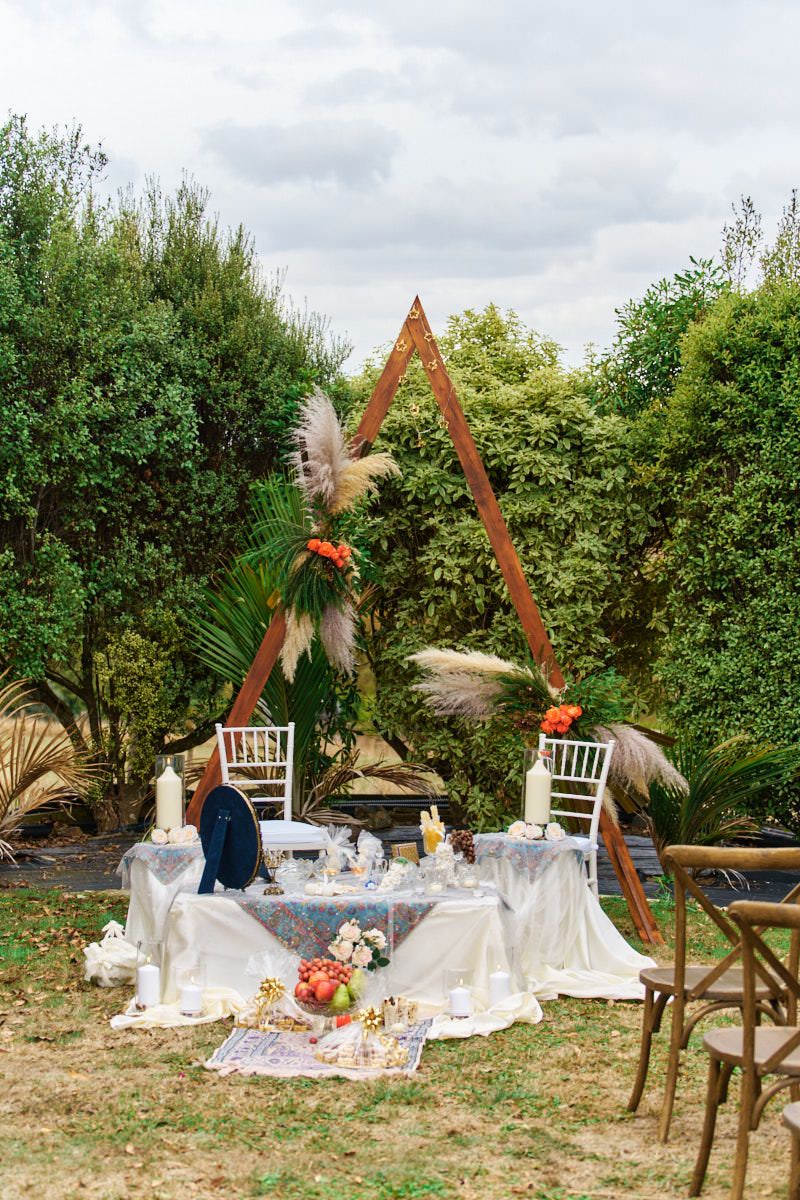 Bohemian outdoor wedding reception table with triangular arch, pampas grass, and floral decor set in a lush garden.