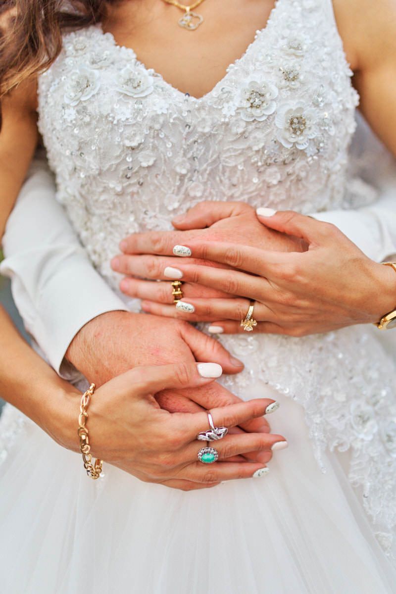 Close-up of bride and groom hands intertwined, adorned with gold rings, bracelets, and jewels against white wedding dress.