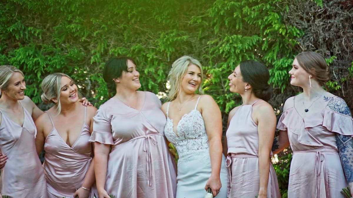 Bride in white gown smiling with five bridesmaids in pink dresses outdoors against lush green foliage.