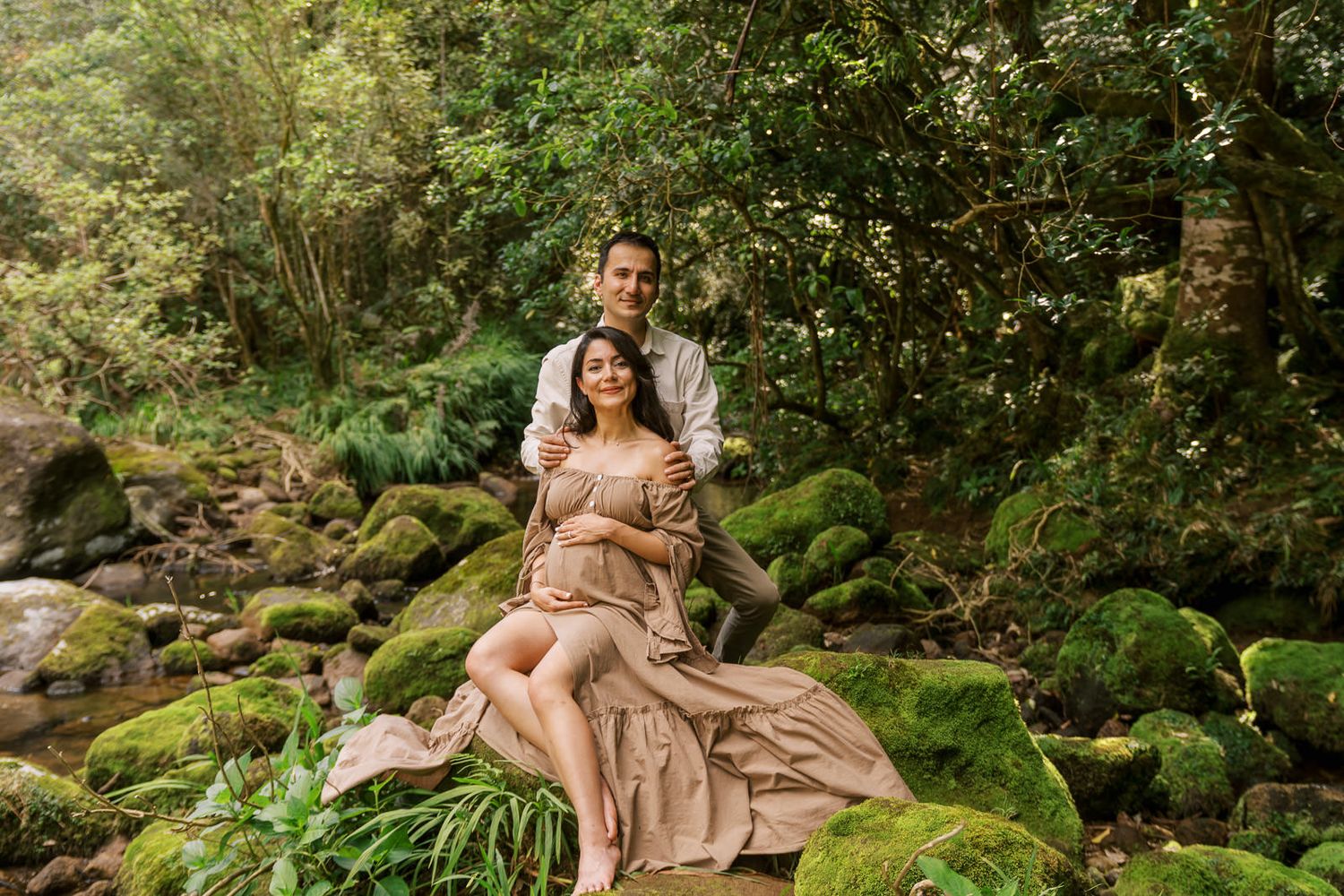 Couple maternity photo shoot in lush forest setting, woman in flowing pink gown seated on moss-covered rocks.