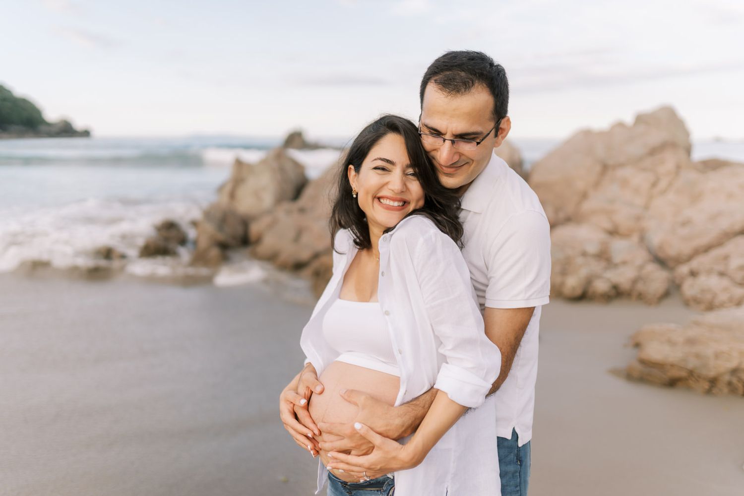 Couple embracing on a beach during a maternity photoshoot, smiling joyfully with rocky coastline in background.