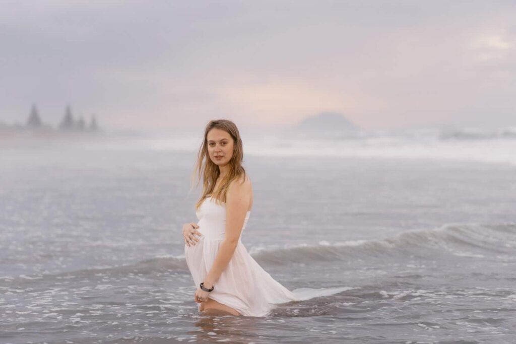 Pregnant woman in white dress standing in ocean waves at sunset during maternity photo shoot.
