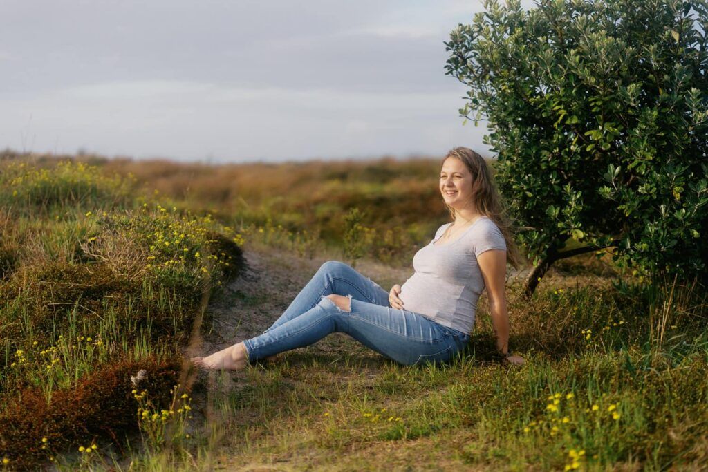 Pregnant woman in white tee and jeans relaxing on grass beside a small tree in a sunny field.
