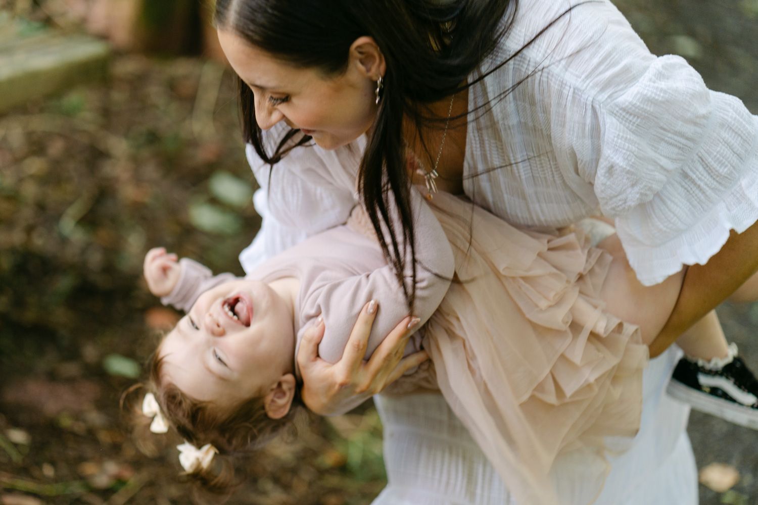 A joyful mother in a white dress playfully holds her laughing toddler upside down outdoors.