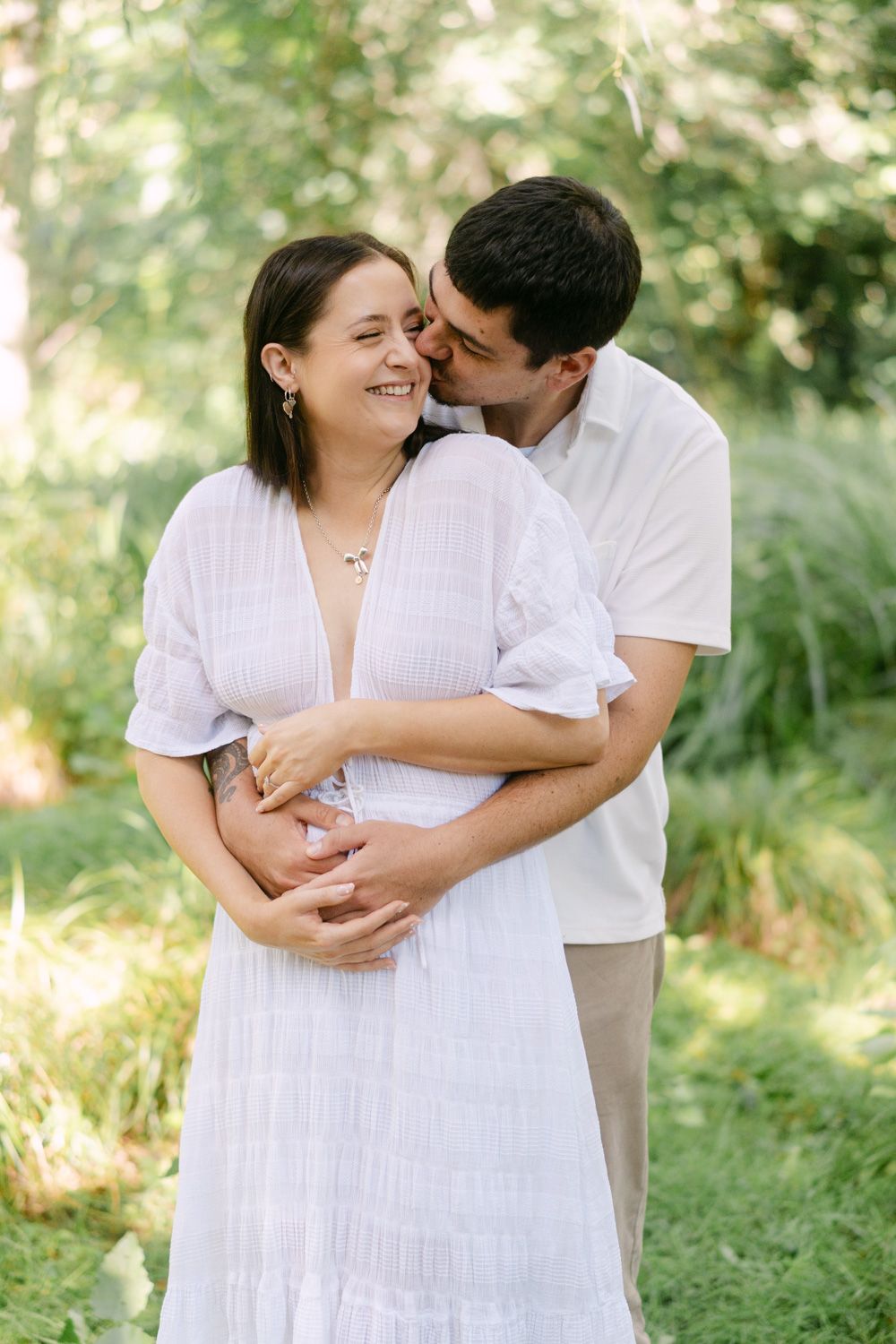 A couple embraces outdoors, woman in white dress smiling as man kisses her cheek, cradling her pregnant belly.
