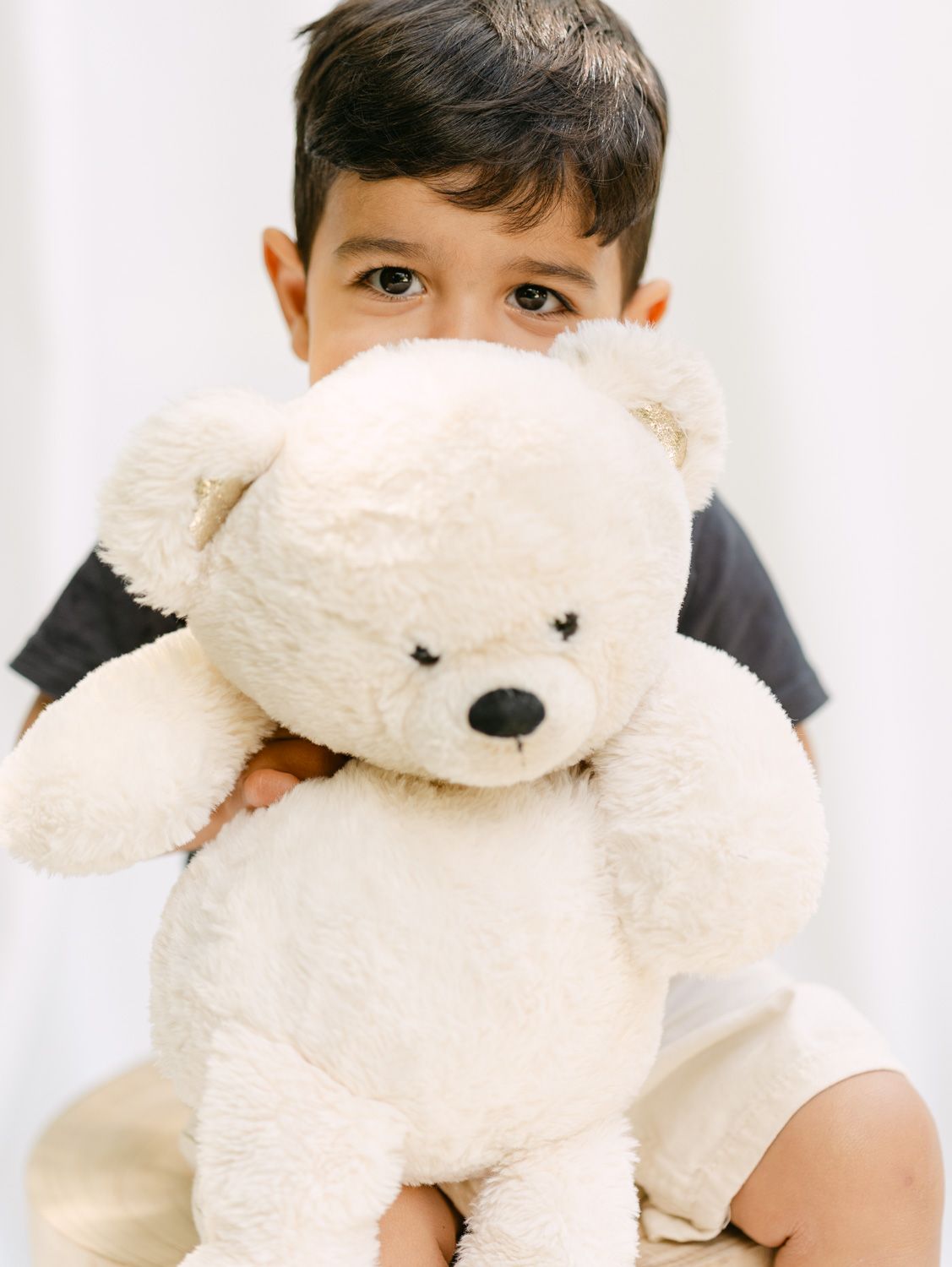 A young boy peeks over a large fluffy white teddy bear, holding it close in a sweet and playful embrace.