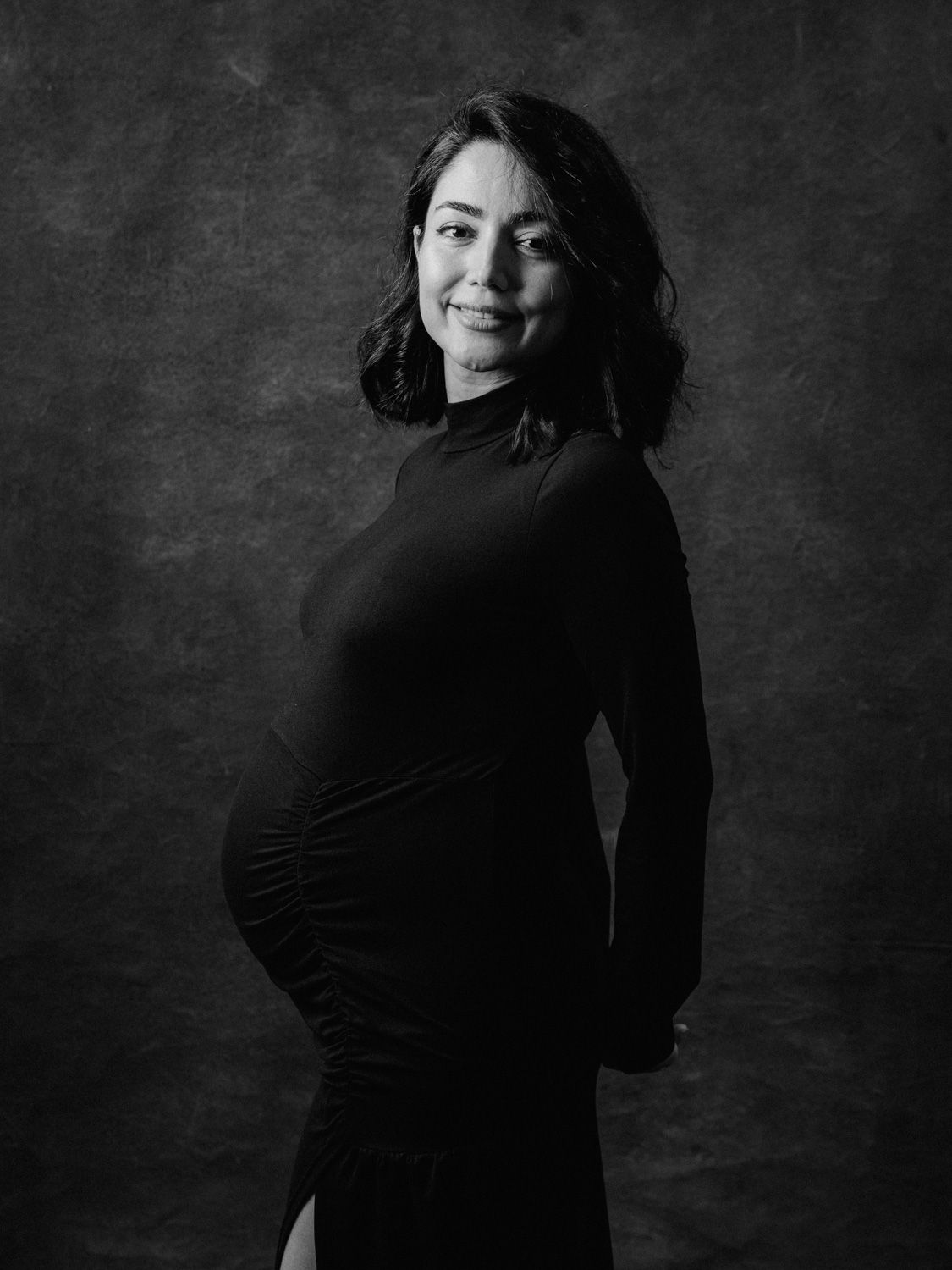Black and white maternity portrait of a smiling woman in a black dress, showcasing her baby bump in a studio setting.