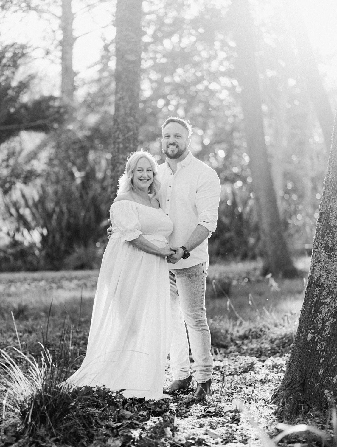 Black and white photo of a couple embracing outdoors among trees, woman in flowing white dress, man in casual white shirt.