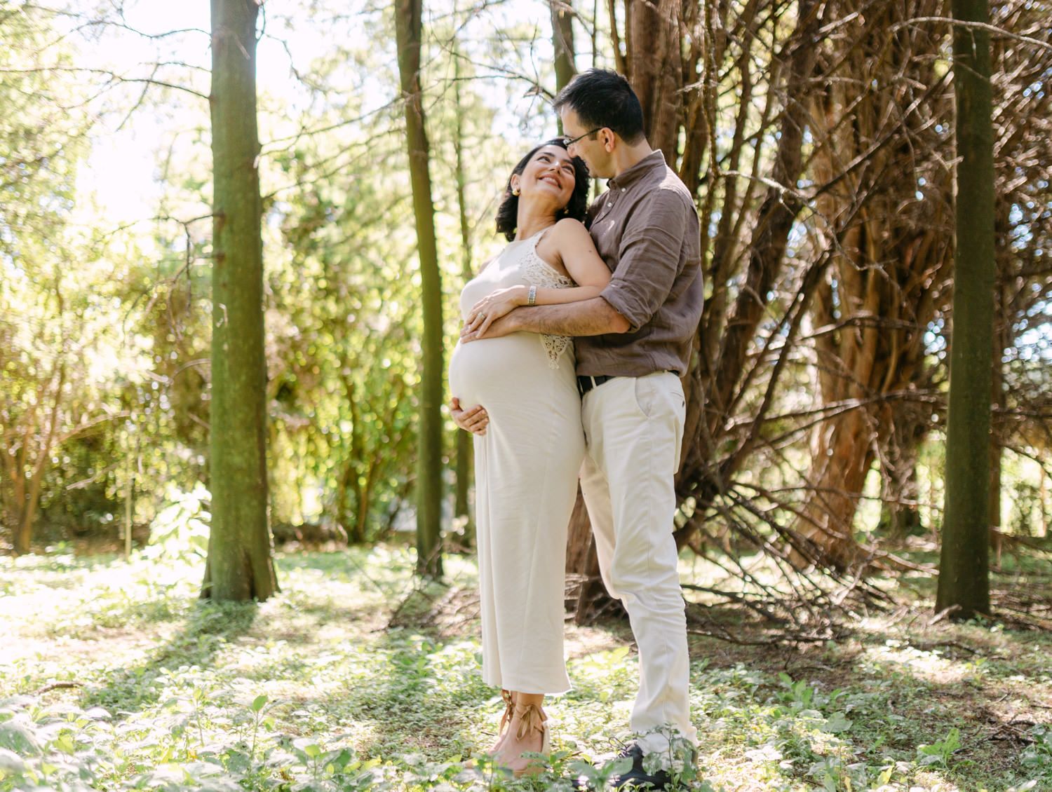 Couple embracing in sunny forest, pregnant woman in white jumpsuit with partner kissing her cheek among trees.