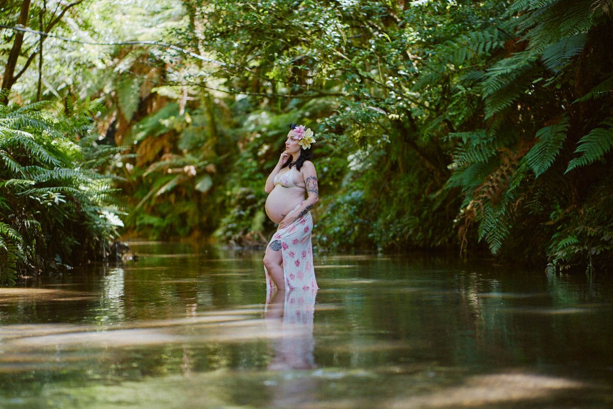 Pregnant woman in floral dress standing in a serene forest stream surrounded by lush green ferns and trees.