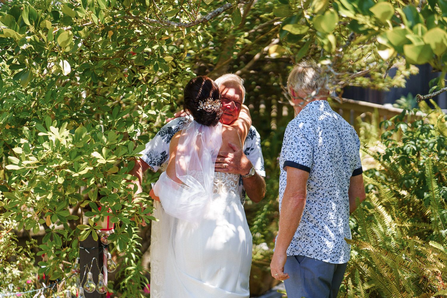 Bride in white gown embracing guests outdoors among lush green trees on a sunny wedding day.