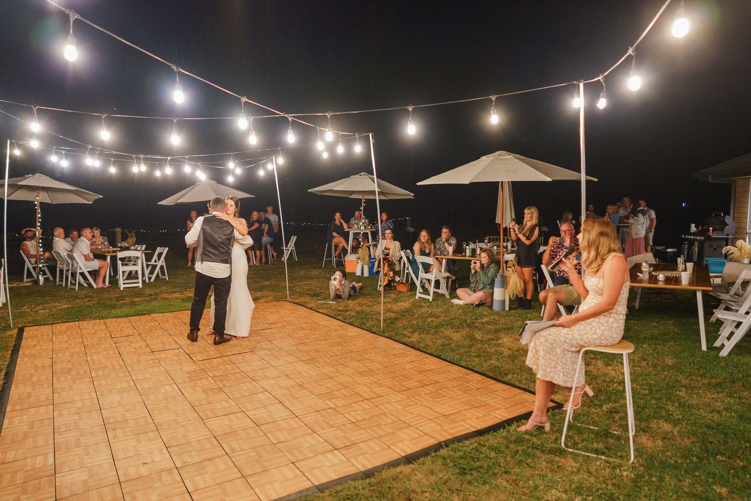 Couple shares first dance at outdoor wedding reception under string lights with guests watching.