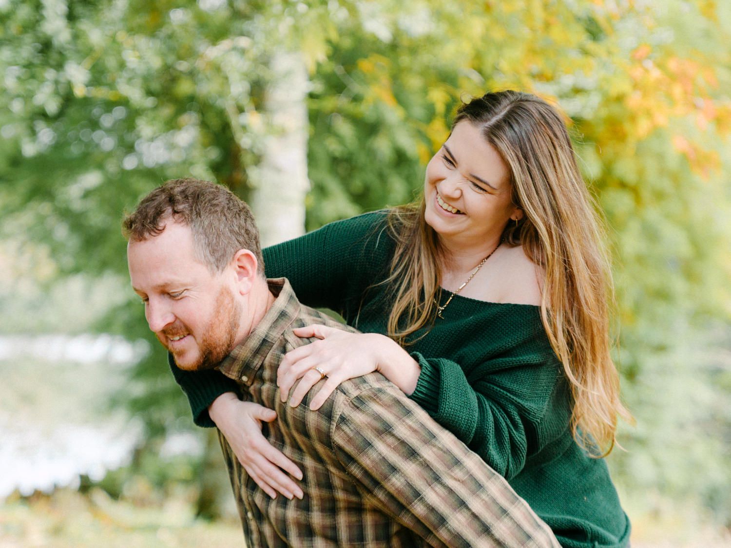 A smiling couple in green outfits share a piggyback ride outdoors among autumn trees.