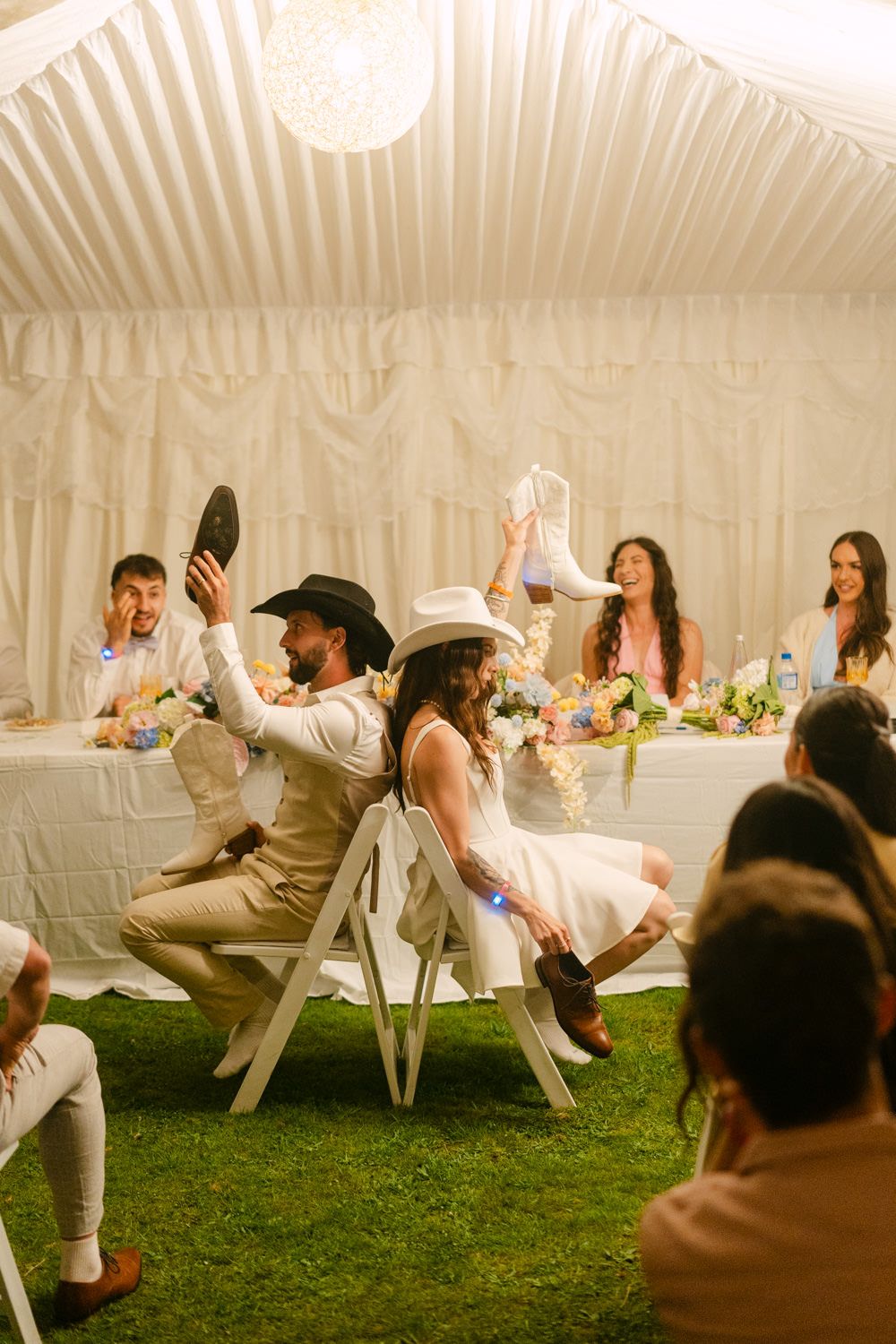 Bride and groom in cowboy hats playfully hold up shoes at rustic outdoor wedding reception under white tent.