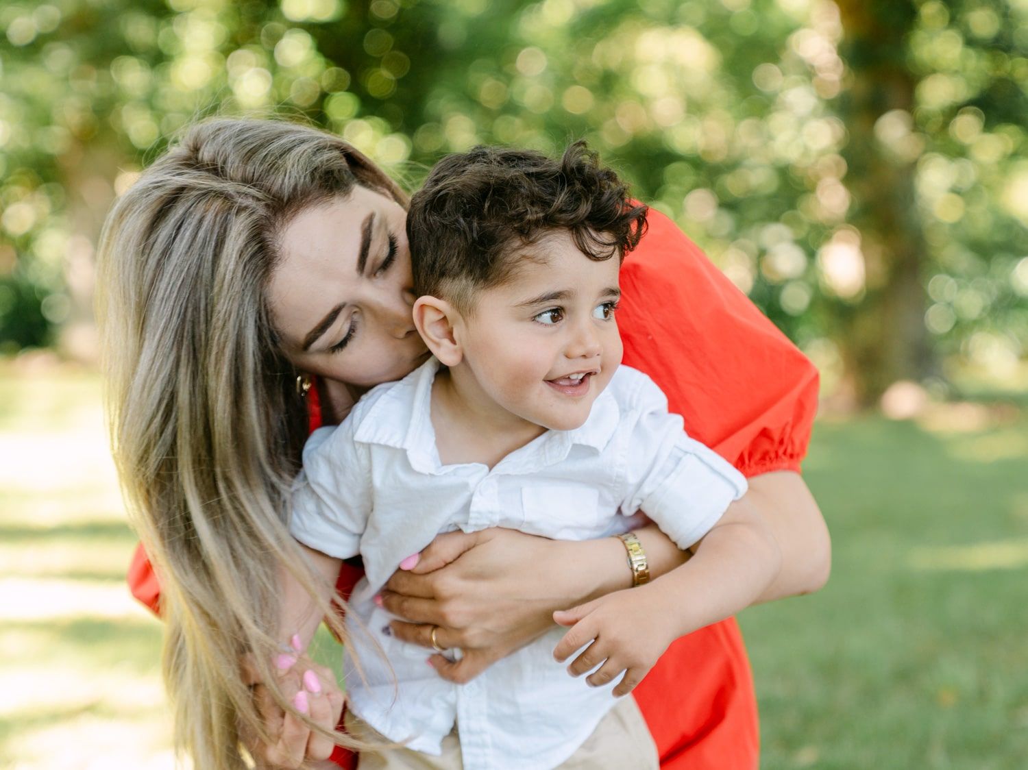 A loving mother in a red dress hugs and kisses her smiling toddler boy outdoors in a lush green park.