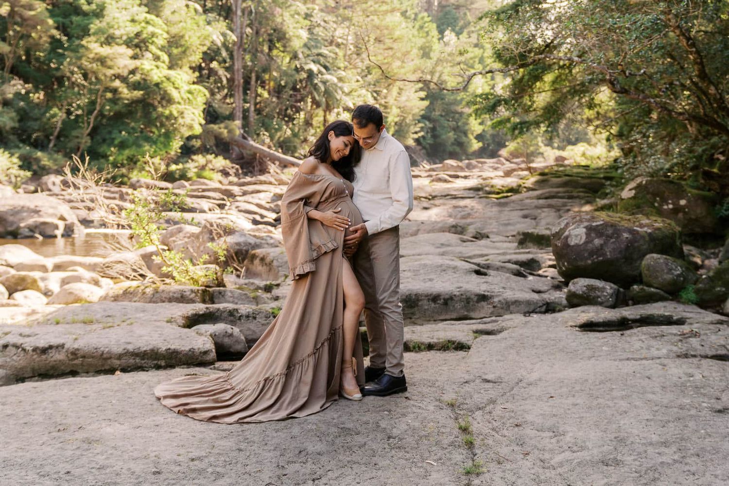 Couple embracing on rocky riverbed surrounded by lush forest, woman in flowing taupe gown, maternity photoshoot.