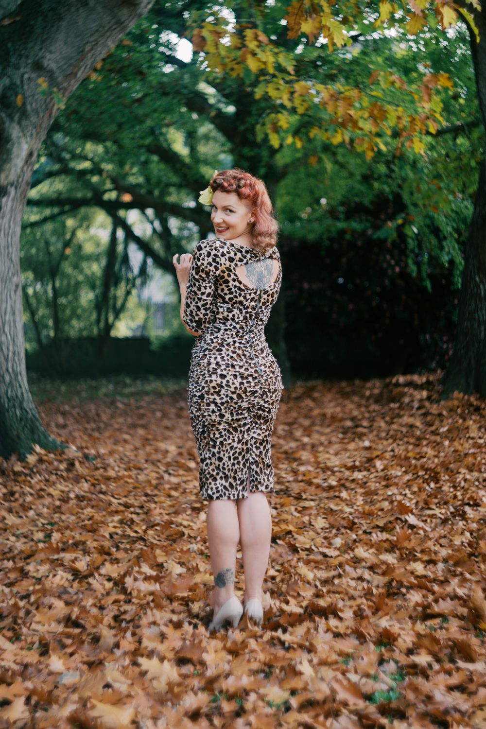Woman in leopard print dress standing among autumn leaves in a tree-lined path.