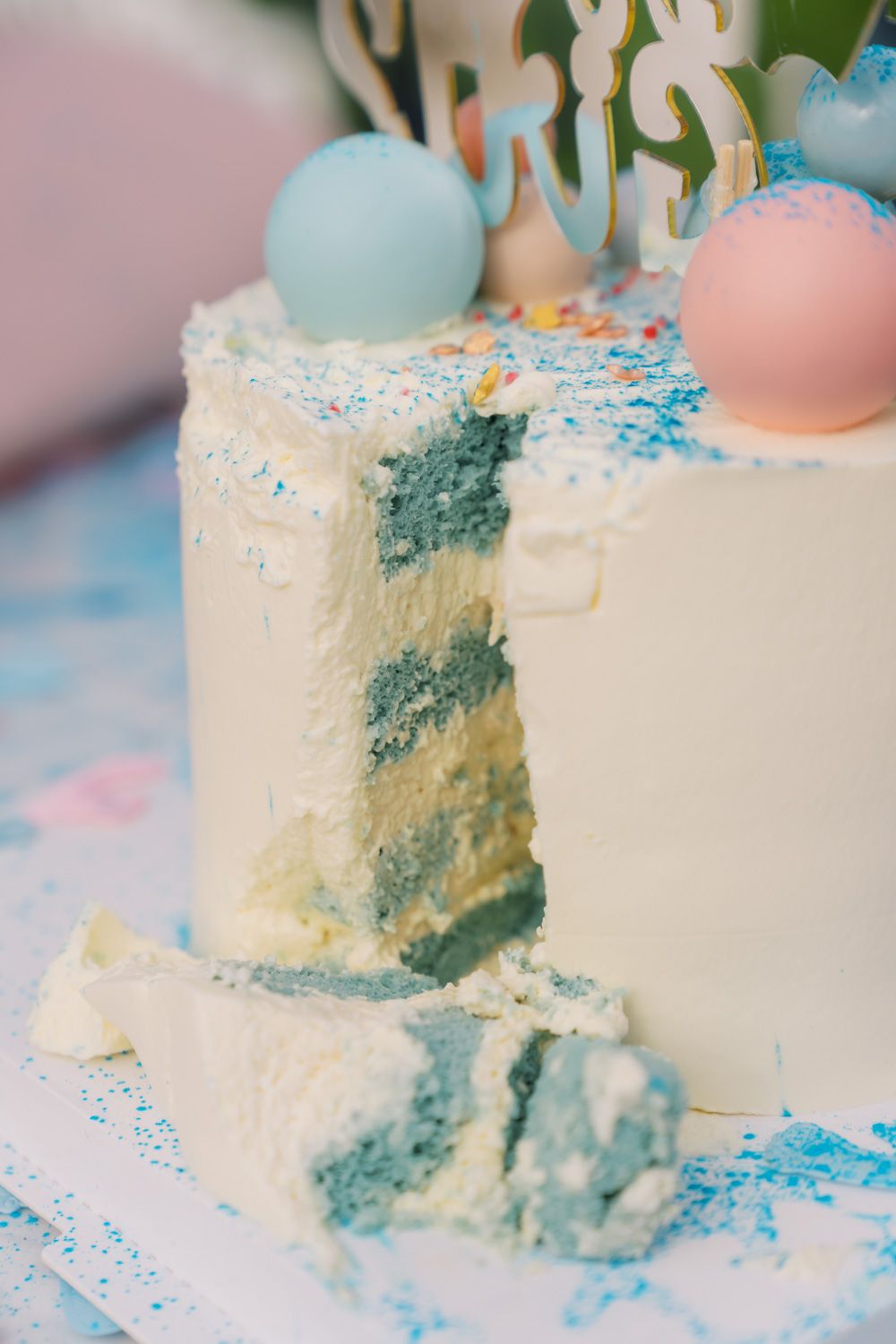 Gender reveal cake with blue interior, white frosting, pastel ball toppers, and blue sprinkles on a decorated table.