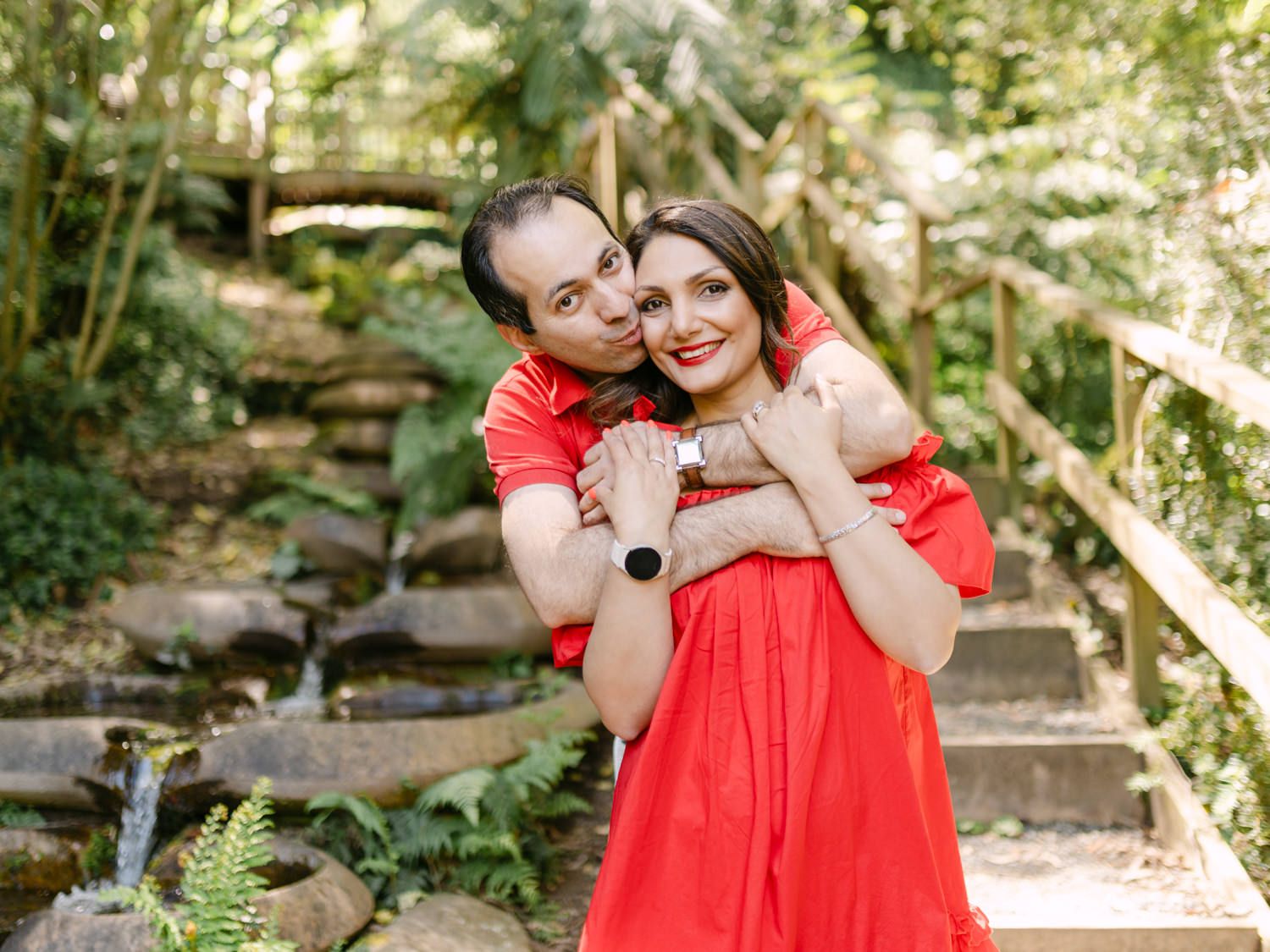 Happy couple in matching red outfits embracing on stone steps surrounded by lush green garden foliage.