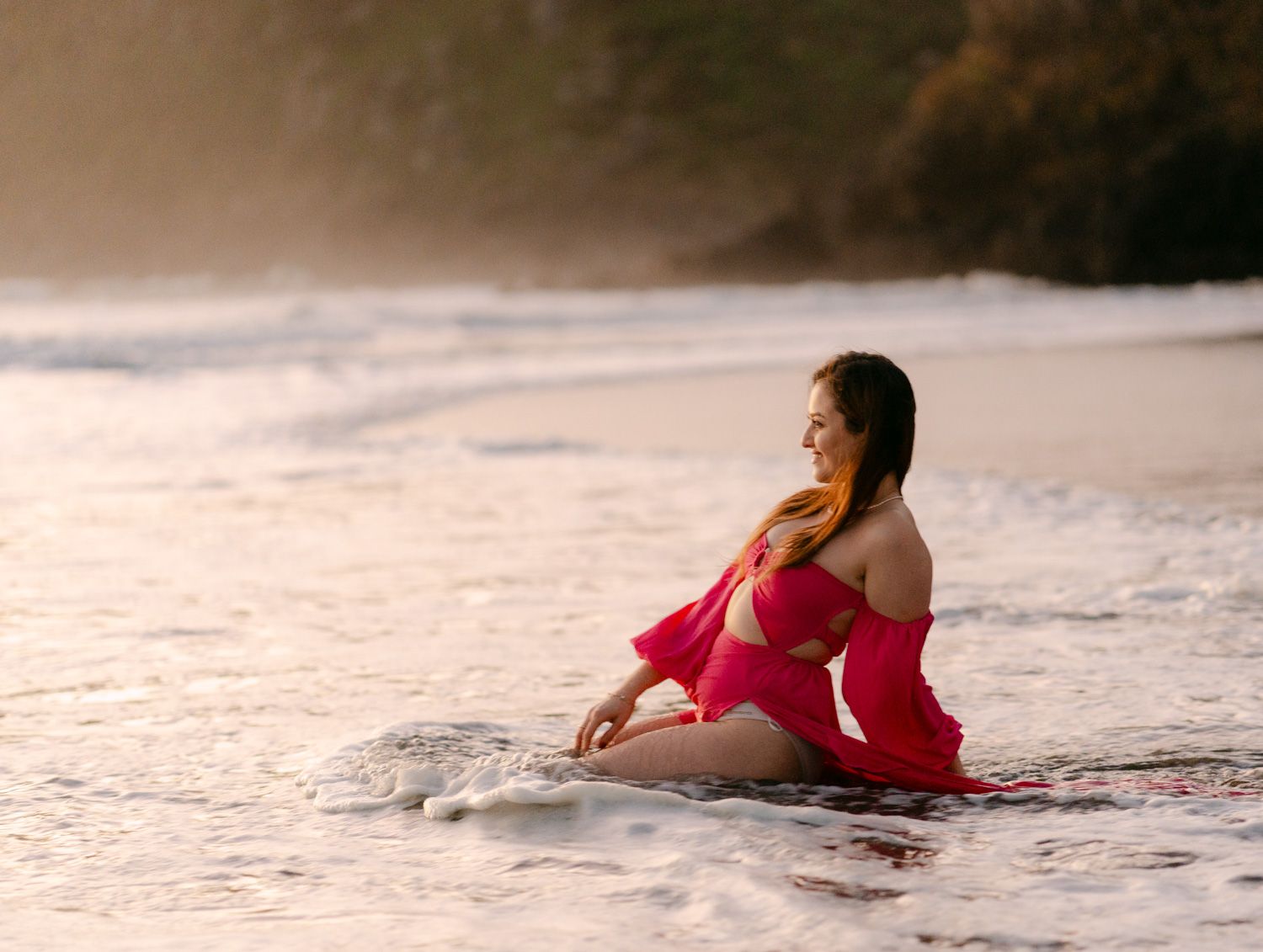 Pregnant woman in red dress sitting on sandy beach with ocean waves at golden hour.