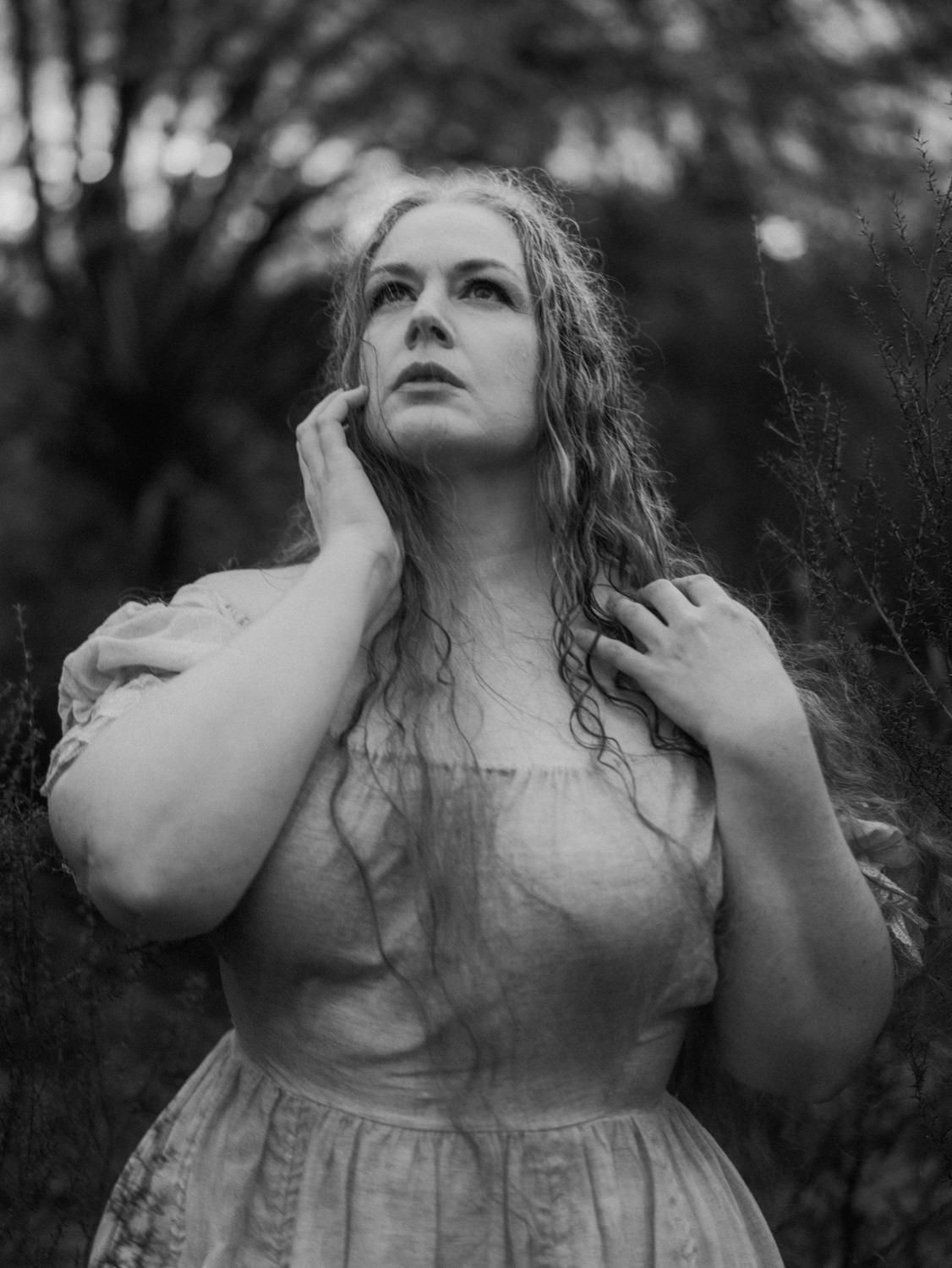 Black and white portrait of a woman in a vintage dress touching her hair outdoors.