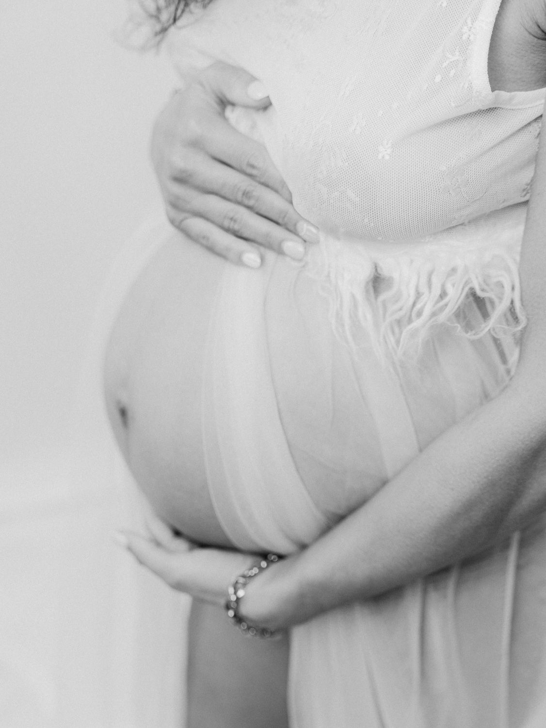 Black and white maternity photo of pregnant woman cradling bare baby bump in flowing lace gown with bracelet.