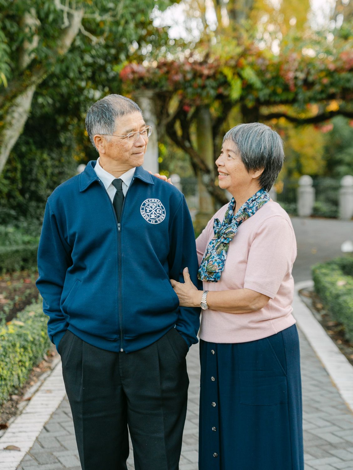 Elderly Asian couple smiling at each other on a garden path, man in blue jacket, woman in pink top.