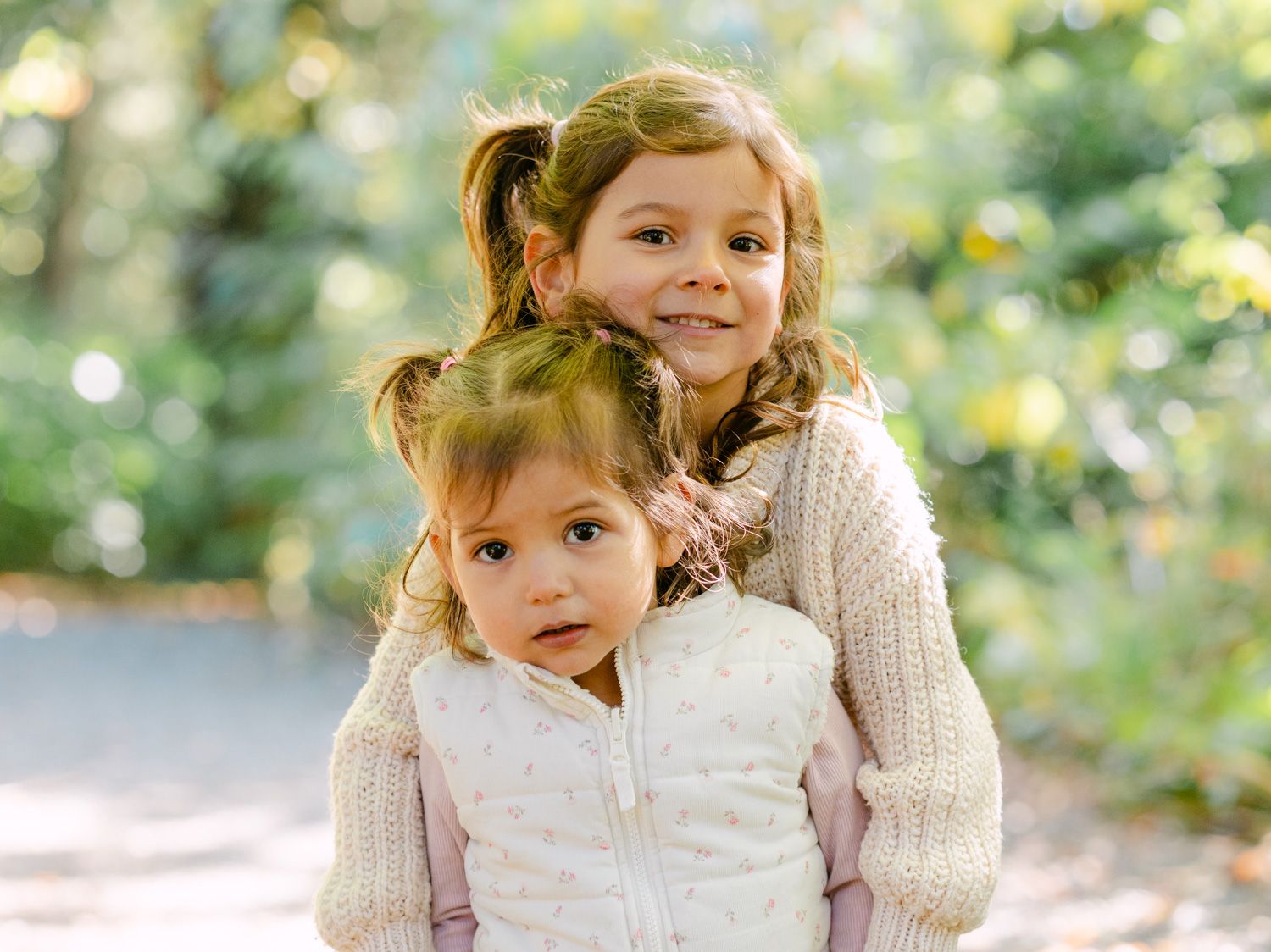Two young sisters pose outdoors in autumn, older girl hugging younger toddler, both wearing cozy cream sweaters.