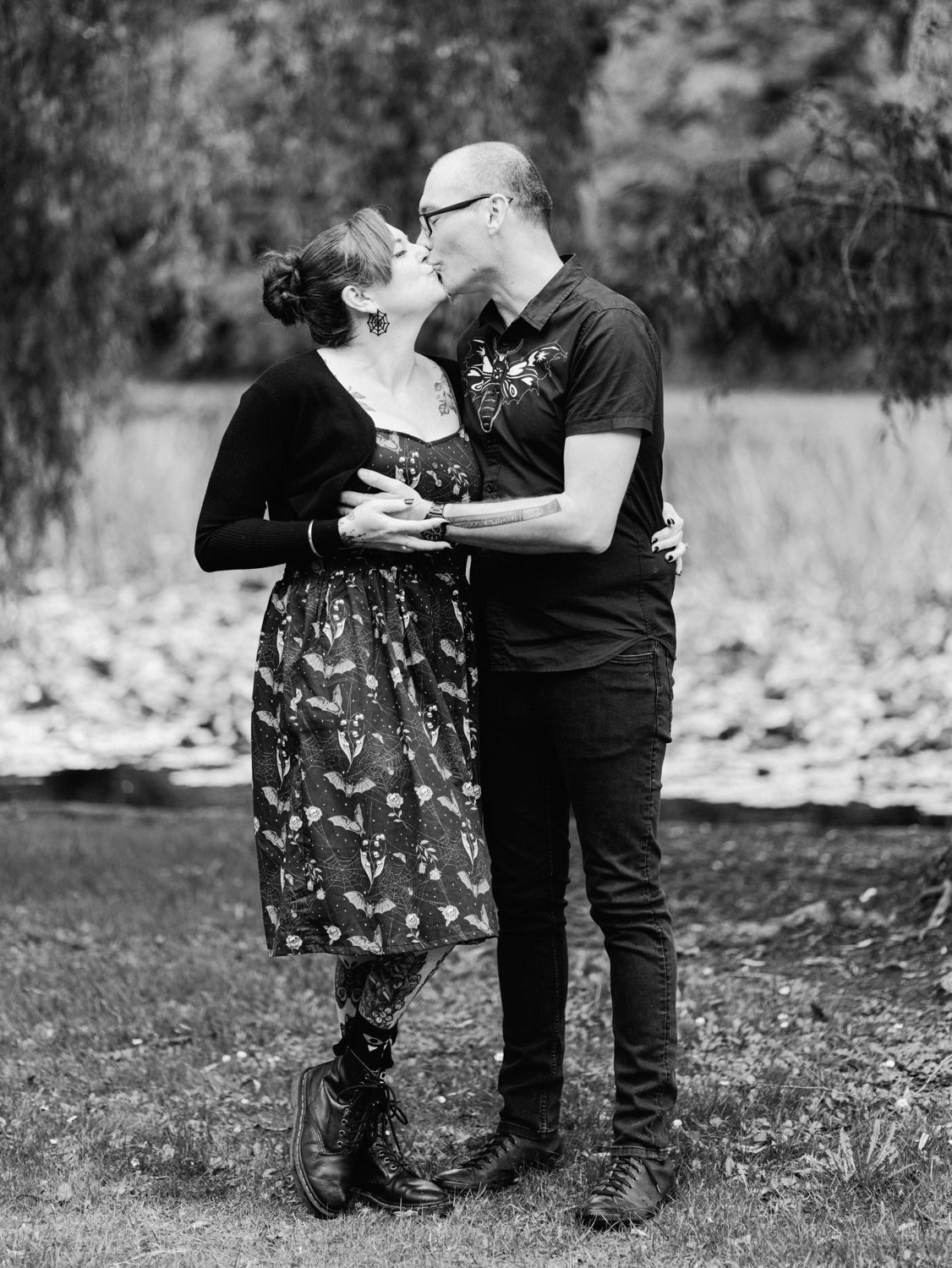 Black and white photo of a couple kissing outdoors near a pond, woman in floral dress and boots, man in black t-shirt.