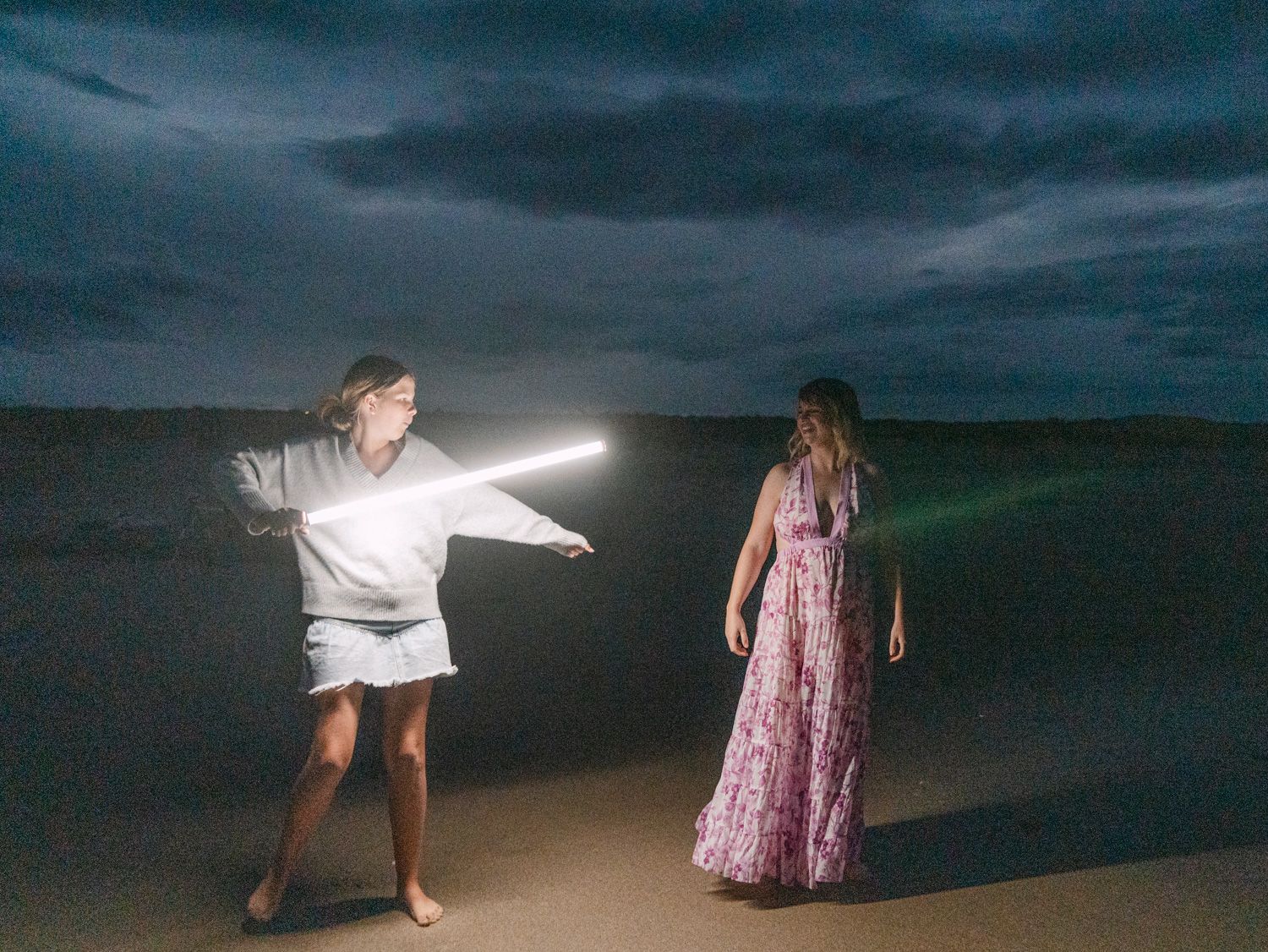 Two women on a beach at dusk, one holding a glowing light tube like a lightsaber in a dramatic scene.