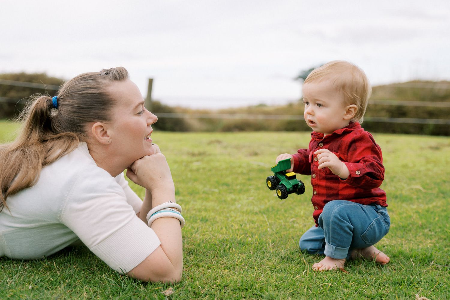 A mother lies on green grass smiling at her toddler boy in a red sweater holding a toy tractor outdoors.