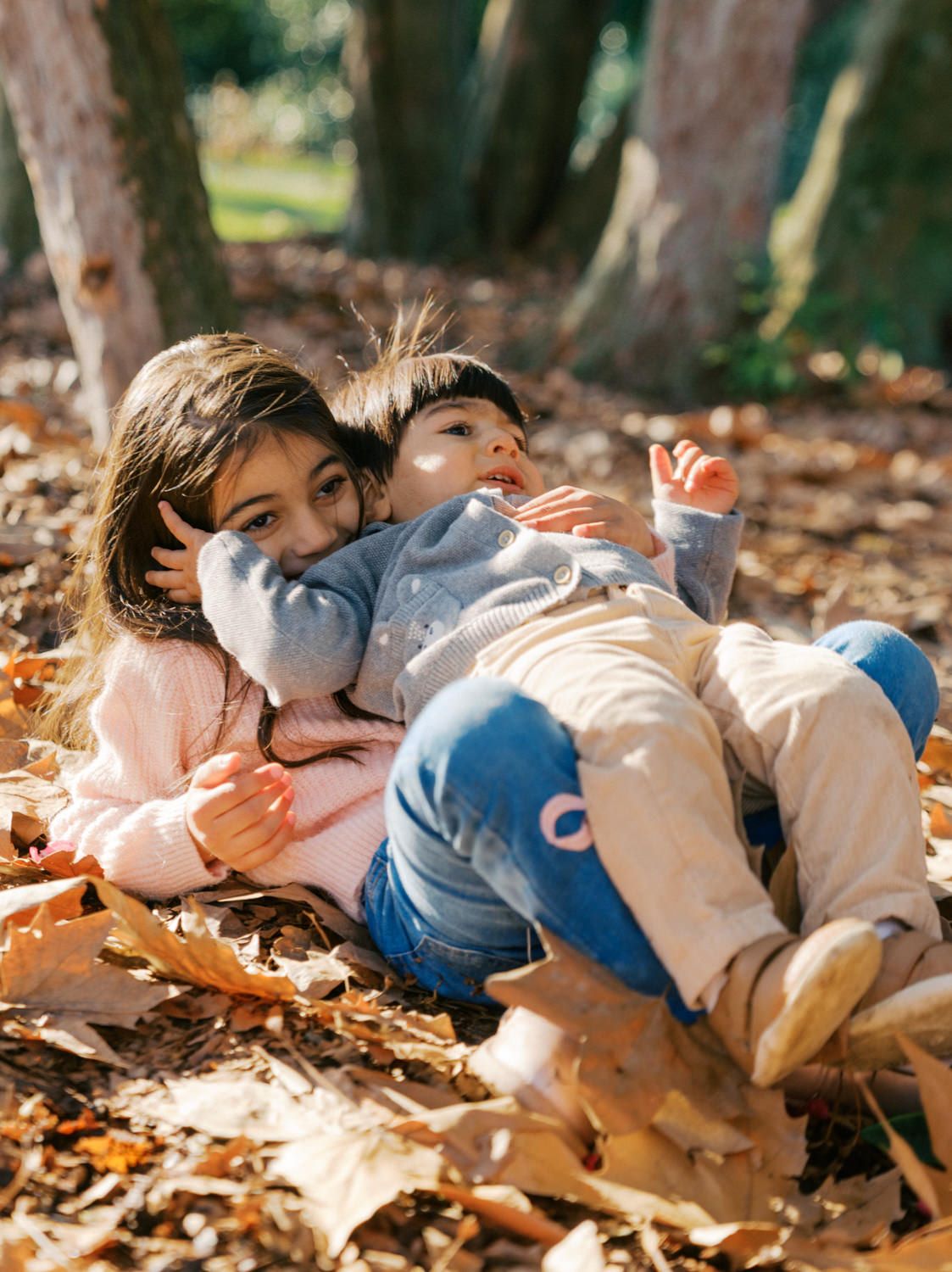 Two children playing in autumn leaves in a sunny park, with the older girl hugging the younger child warmly.