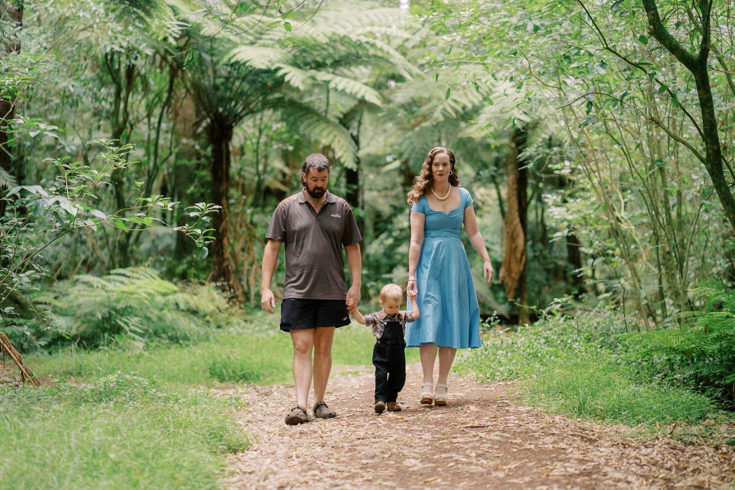 Family of three walking together on a dirt path through a lush green forest.