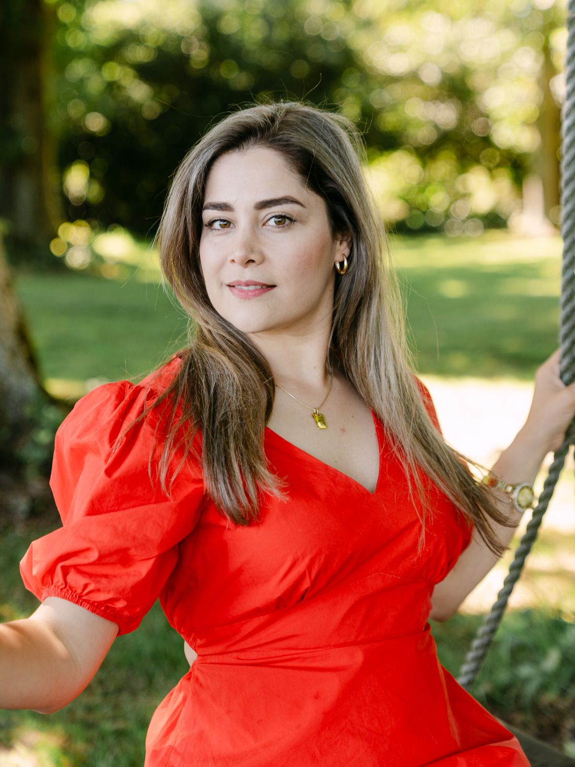 Woman in red puff-sleeve dress sitting on a rope swing outdoors, surrounded by lush green trees.