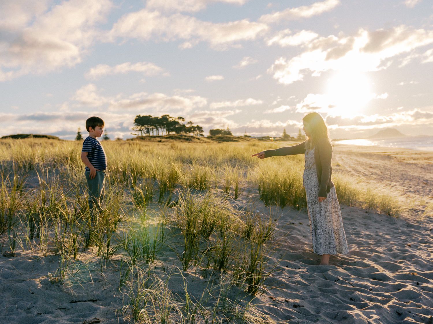 Two children stand on a sandy beach at golden sunset, surrounded by tall grass dunes and a glowing coastal horizon.