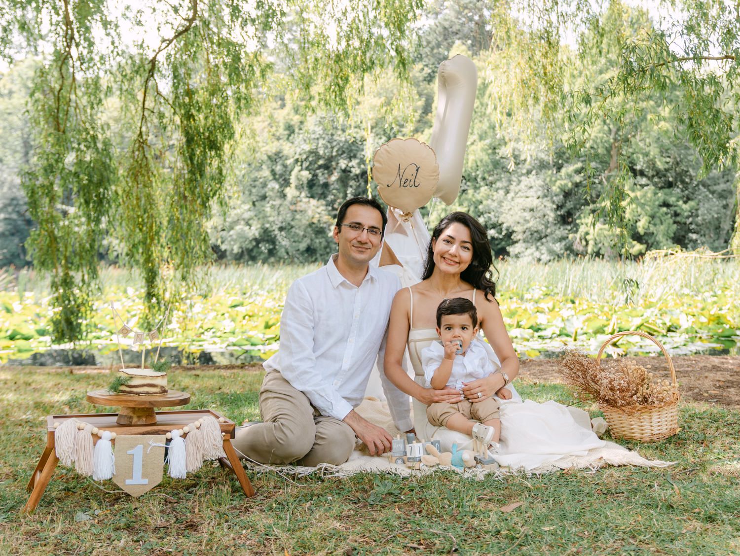 Family of three posing on a picnic blanket outdoors under a willow tree with a balloon and basket nearby.