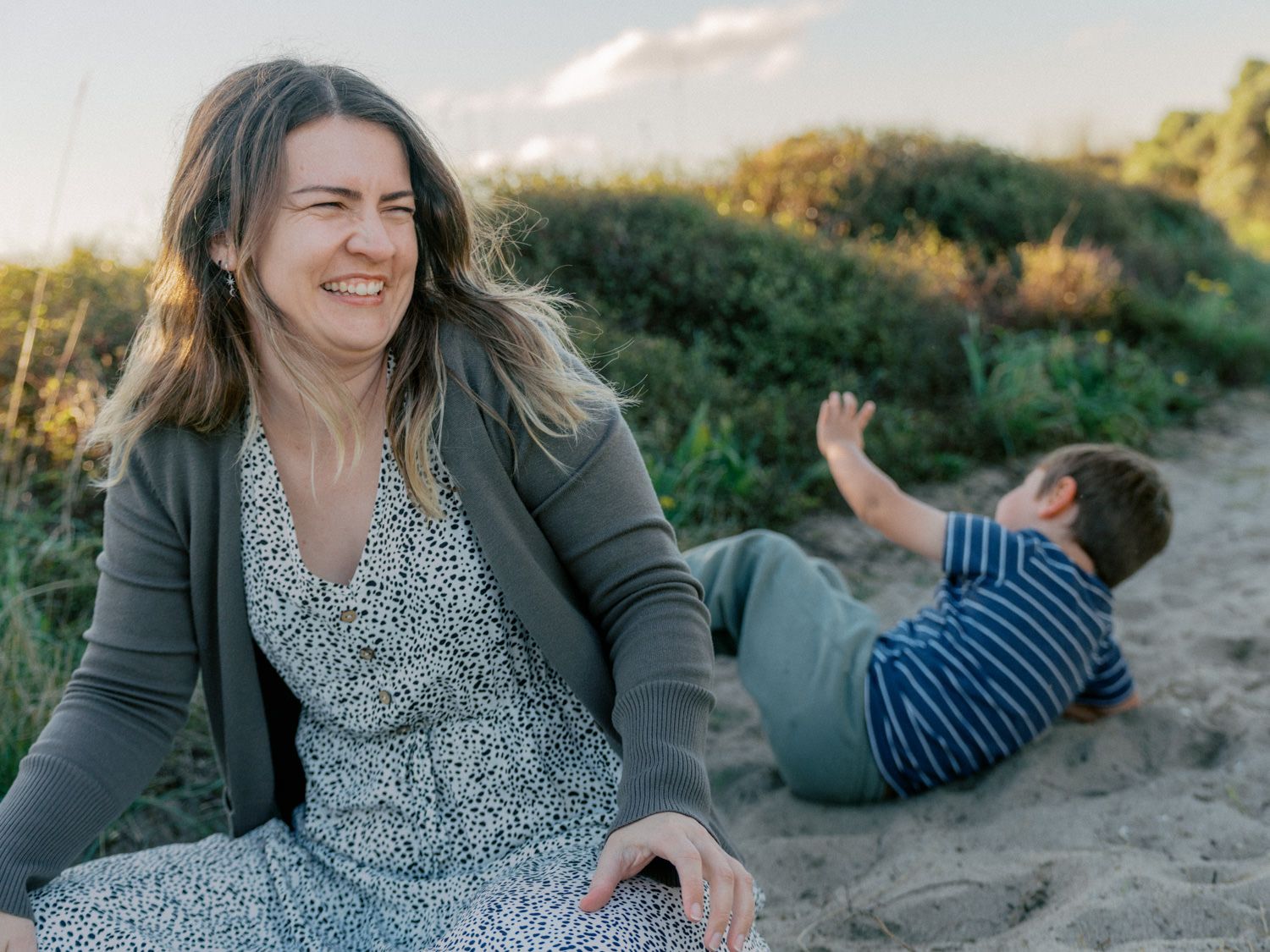 A smiling woman in a floral dress sits on sandy dunes while a child plays behind her at golden hour.