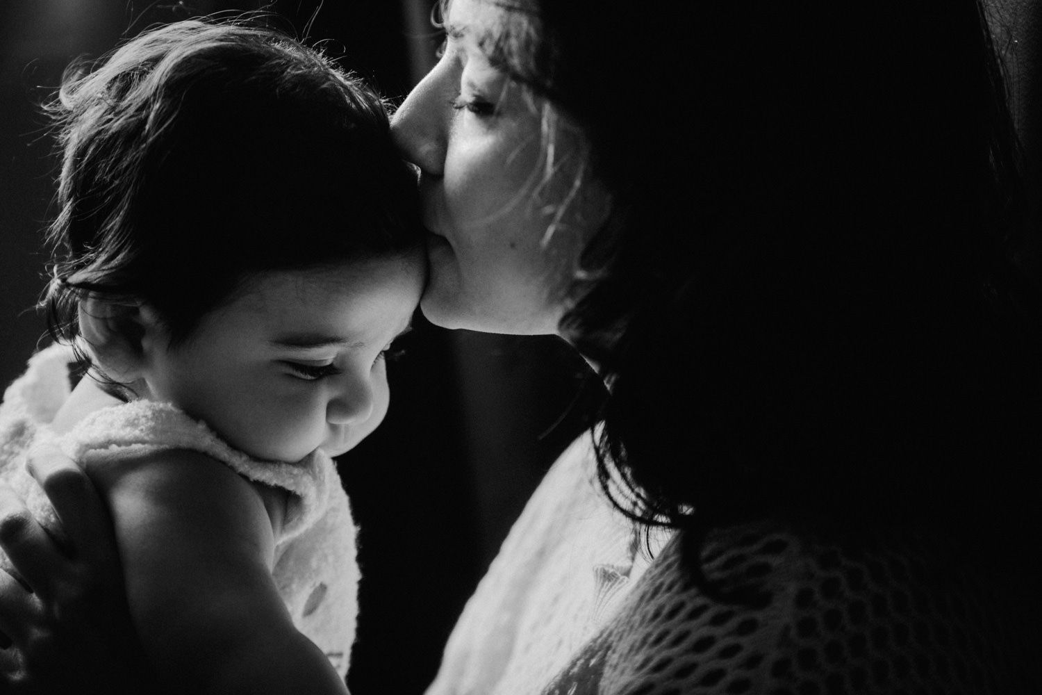 Black and white photo of a mother tenderly kissing her smiling baby on the forehead in soft light.